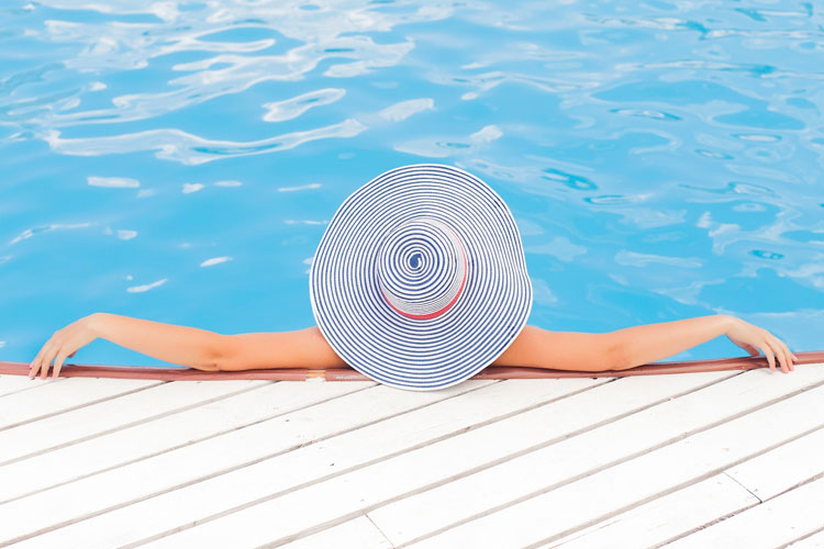 [fotografía] sombrero femenino de anchas alas en primer plano, que oculta una mujer recostada sobre el bordillo de una piscina con el cuerpo en el agua