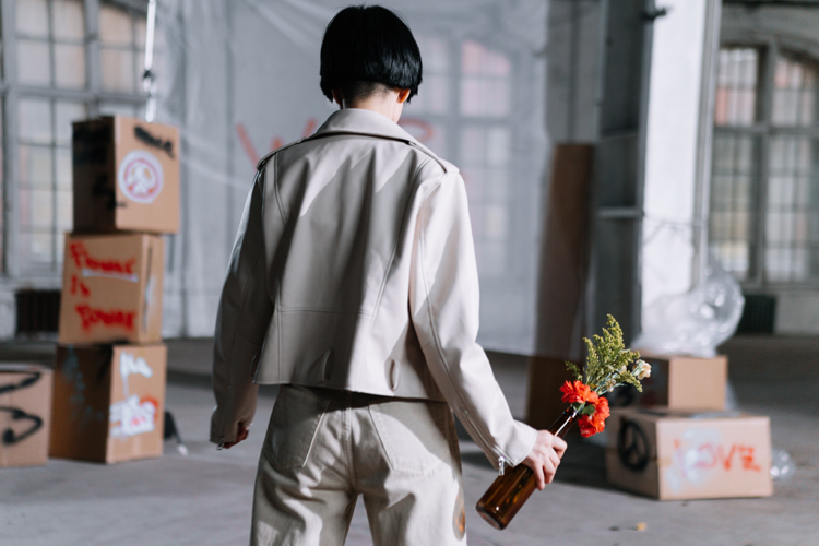 [fotografía] Varón de espaldas en una nave que podría ser un estudio de artista, con una botella en la mano de la que salen flores, al estilo del conocido cóctel molotov de la obra urbana de Banksy
