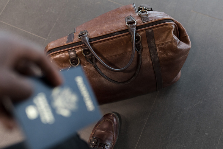 [fotografía] imagen desenfocada de una mano sosteniendo un pasaporte en primer plano, en picado sobre el suelo, donde se ve un zapato y una bolsa de viaje