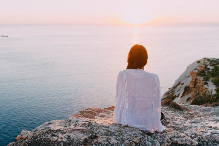 [fotografía] mujer solitaria sentada de espaldas en un acantilado, contemplando una puesta de sol sobre el mar
