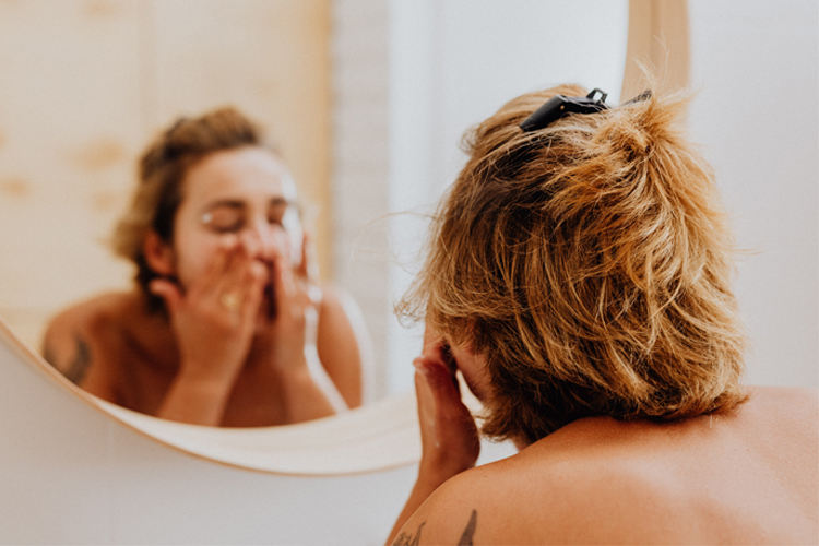 [fotografía] mujer masajeándose la cara con ambas manos delante de un espejo