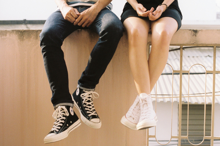 [fotografía] extremidades inferiores de varón y mujer sentados en una tapia baja, él con las piernas abiertas